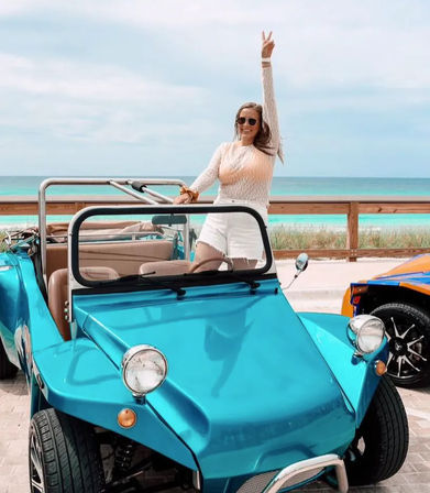 Person in sunglasses standing in a bright turquoise dune buggy on a coastal boardwalk, flashing a peace sign with the beach and turquoise ocean behind