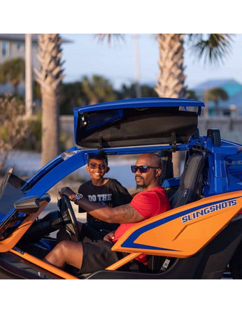 Dad and son grinning in a sporty blue-and-orange three-wheeled roadster parked on a palm-lined coastal road at sunset