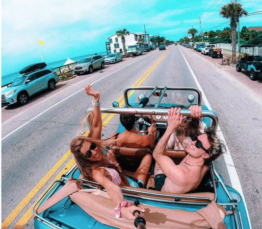 Group of friends in a turquoise convertible cruising down a sunny, palm-lined coastal road with parked cars and the ocean visible on the left — beach road summer road trip vibe.