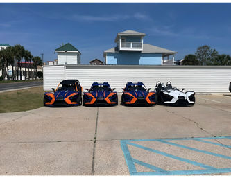 Row of four Polaris Slingshot three-wheeled roadsters (three blue-orange, one white) parked in a coastal beach-town lot with palm trees and pastel beach houses