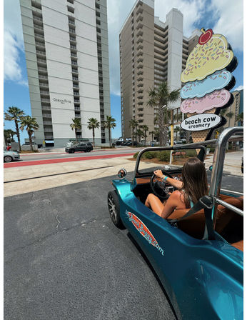 Woman driving a teal beach buggy past a colorful oversized ice cream shop sign and tall oceanfront condominium towers on a sunny coastal street