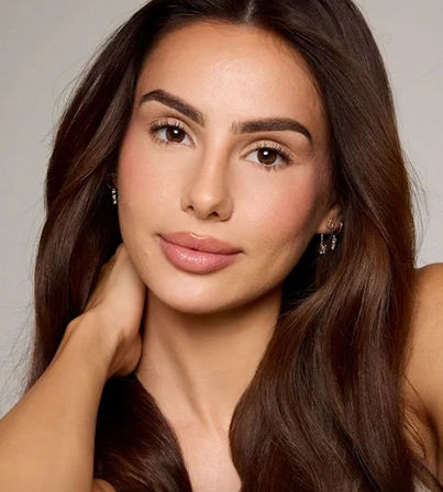 Close-up studio headshot of a woman with long brown hair, clear skin, natural makeup, defined brows, glossy lips and small earrings — warm beauty portrait.