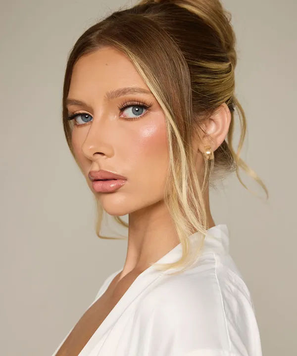 Studio beauty portrait of a woman with a sleek blonde updo, loose face‑framing strands, glowing bridal makeup, blue eyes and a pearl stud earring against a neutral background
