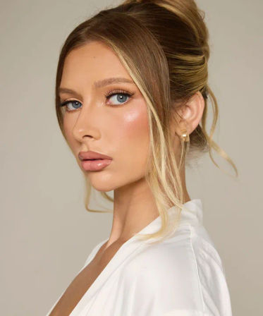 Studio beauty portrait of a woman with a sleek blonde updo, loose face‑framing strands, glowing bridal makeup, blue eyes and a pearl stud earring against a neutral background