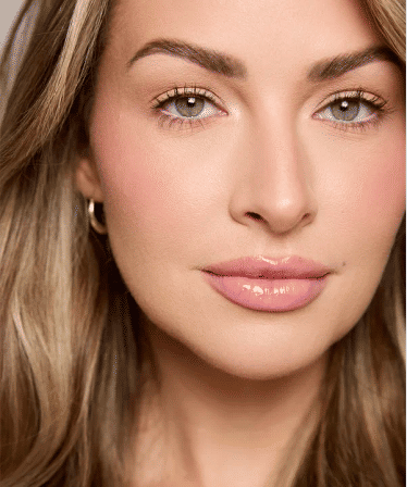 Close-up beauty portrait of a woman with green eyes, arched brows, glossy pink lips, subtle blush, long blonde hair and a small gold hoop earring.