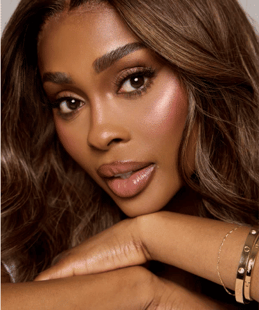 Glam beauty close-up of a woman with glowing skin, shimmery pink blush, glossy nude lips, long lashes and wavy brown hair, chin resting on crossed arms wearing gold bracelets, studio makeup portrait
