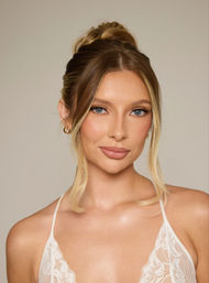 Indoor studio headshot of a woman with a blonde high bun, face‑framing tendrils, blue eyes and natural makeup, glowing skin, wearing a white lace camisole against a neutral background