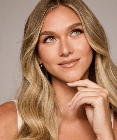 Studio beauty headshot of a smiling blonde woman with long wavy hair and natural makeup, hand under chin and looking upward