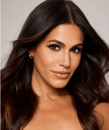 Close-up studio headshot of a woman with long dark hair, bronzed glowing skin, defined brows and lashes, nude glossy lips and a cheek beauty mark — glamorous beauty makeup portrait.
