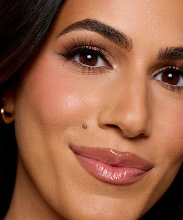 Close-up beauty portrait of a woman with arched brows, long fluttery lashes, warm bronzy eyeshadow, dewy medium-toned skin, glossy nude lips and a small cheek mole.