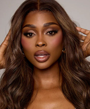 Close-up beauty portrait of a woman with long wavy brown hair and glam smokey-eye makeup, rosy cheeks, glossy nude lips and gold hoop earring, hands framing her face in a studio headshot.