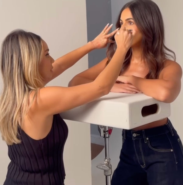 Behind-the-scenes makeup artist applying under-eye makeup to a model leaning on a white prop box in a bright photo studio