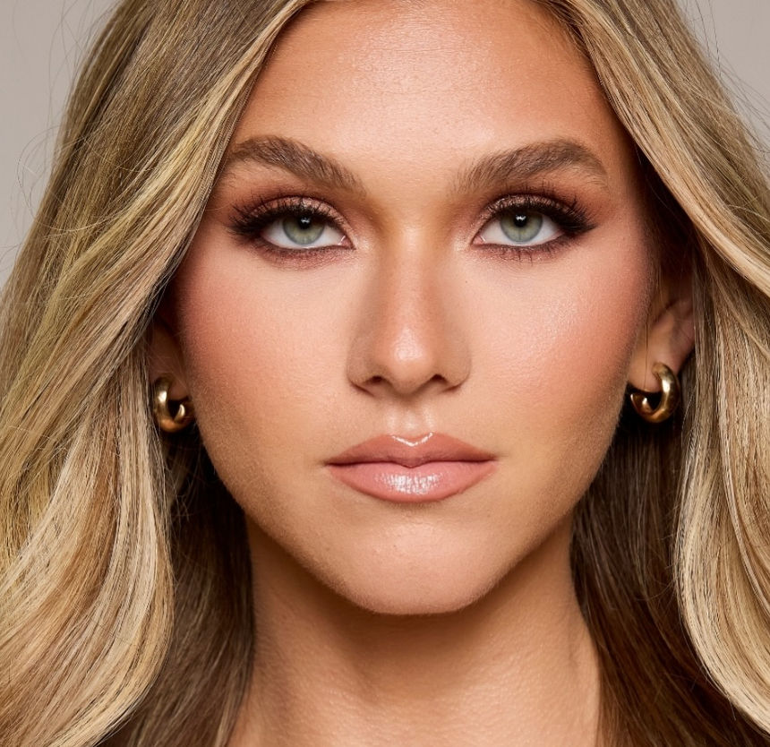 Close-up beauty portrait of a blonde woman with green eyes, glossy nude lips, smoky natural-glam makeup and gold hoop earrings in a studio-lit cosmetic headshot