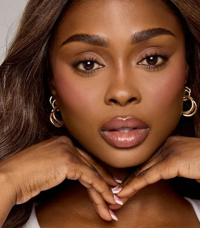 Close-up beauty portrait of a woman with glowing skin, glossy lips, sculpted brows and gold hoop earrings, hands framing her chin — studio makeup and lighting cosmetic shot.