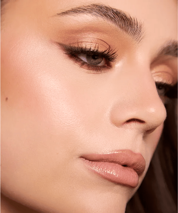 Glam beauty close-up: shimmery gold eyeshadow, soft winged eyeliner, long lashes, glossy nude lips and dewy highlighted skin.