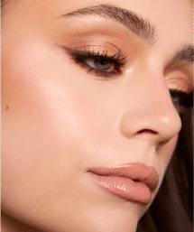 Glam beauty close-up: shimmery gold eyeshadow, soft winged eyeliner, long lashes, glossy nude lips and dewy highlighted skin.