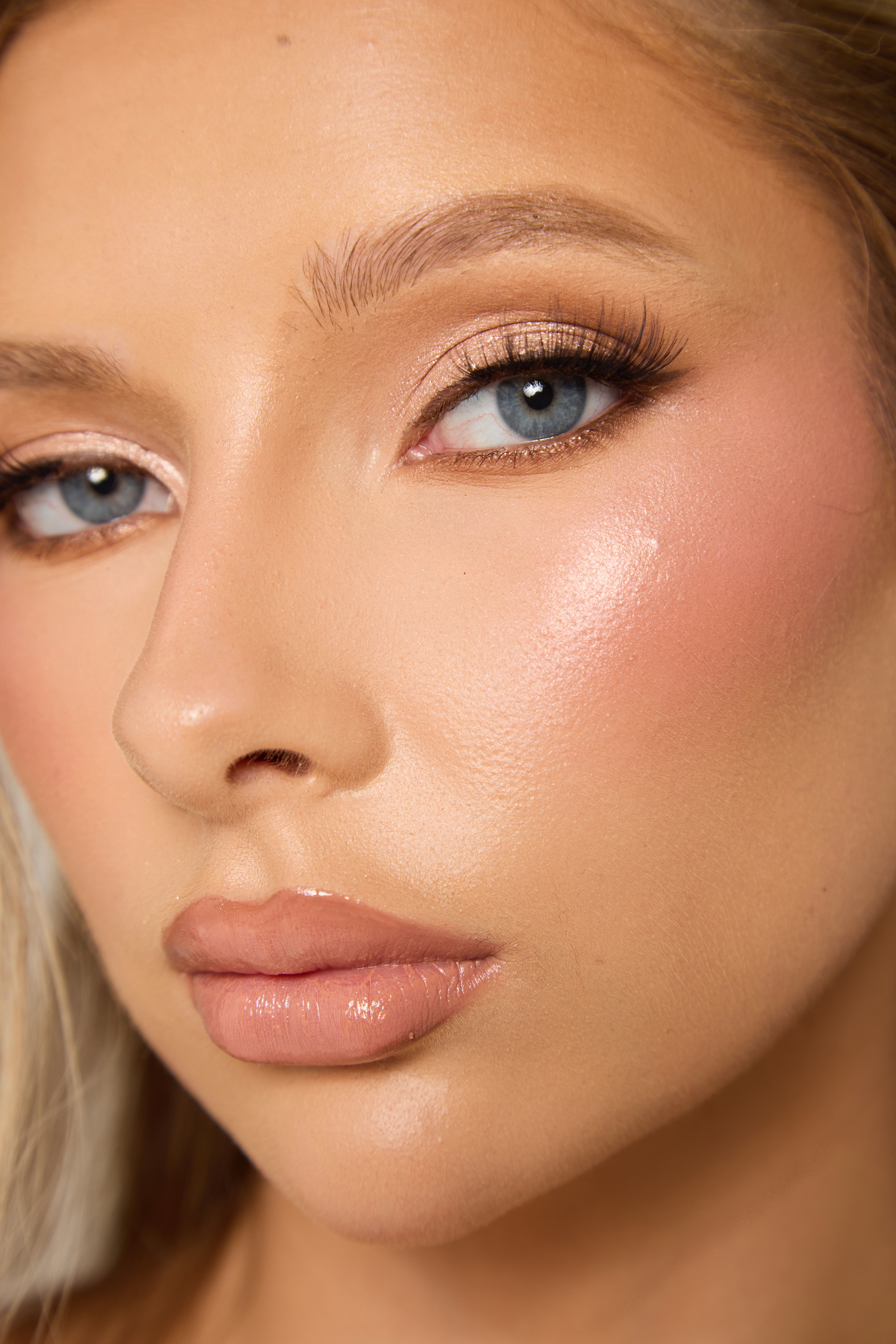Close-up beauty portrait of a face with blue eyes, shimmery gold eyeshadow, long lashes, glowing dewy skin, rosy highlight and glossy nude lips.