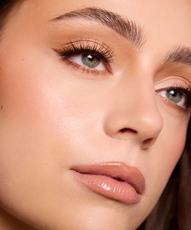 Close-up beauty portrait of a face with green eyes, long lashes, full brows, dewy flawless skin and glossy nude lips — natural makeup look.
