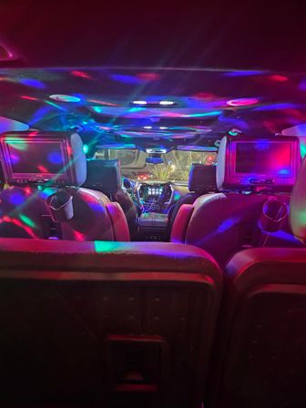 Backseat view inside an SUV at night with colorful red, blue and green disco lights reflecting across leather seats, dashboard touchscreen and dual headrest entertainment screens, playful party-style car interior.
