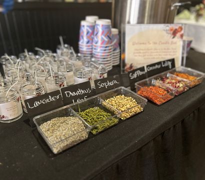 Close-up of an event DIY station with handmade jar candles and wicks, chalkboard labels (lavender), clear trays of dried herbs and colorful safflower petals, and stacked paper cups on a black tablecloth.