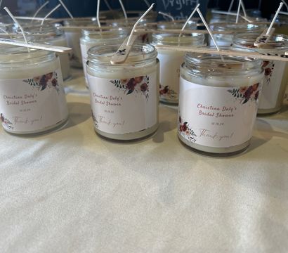 Rows of glass jar candles with wooden stick wick holders and floral personalized labels, arranged as bridal shower favors on a cream tablecloth