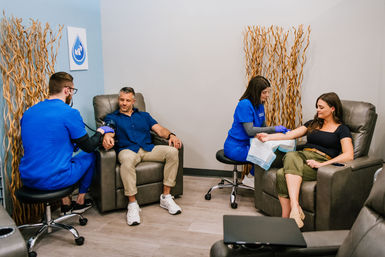 Adults receiving IV therapy in a wellness clinic lounge, medical staff in blue scrubs administering treatments in reclining chairs