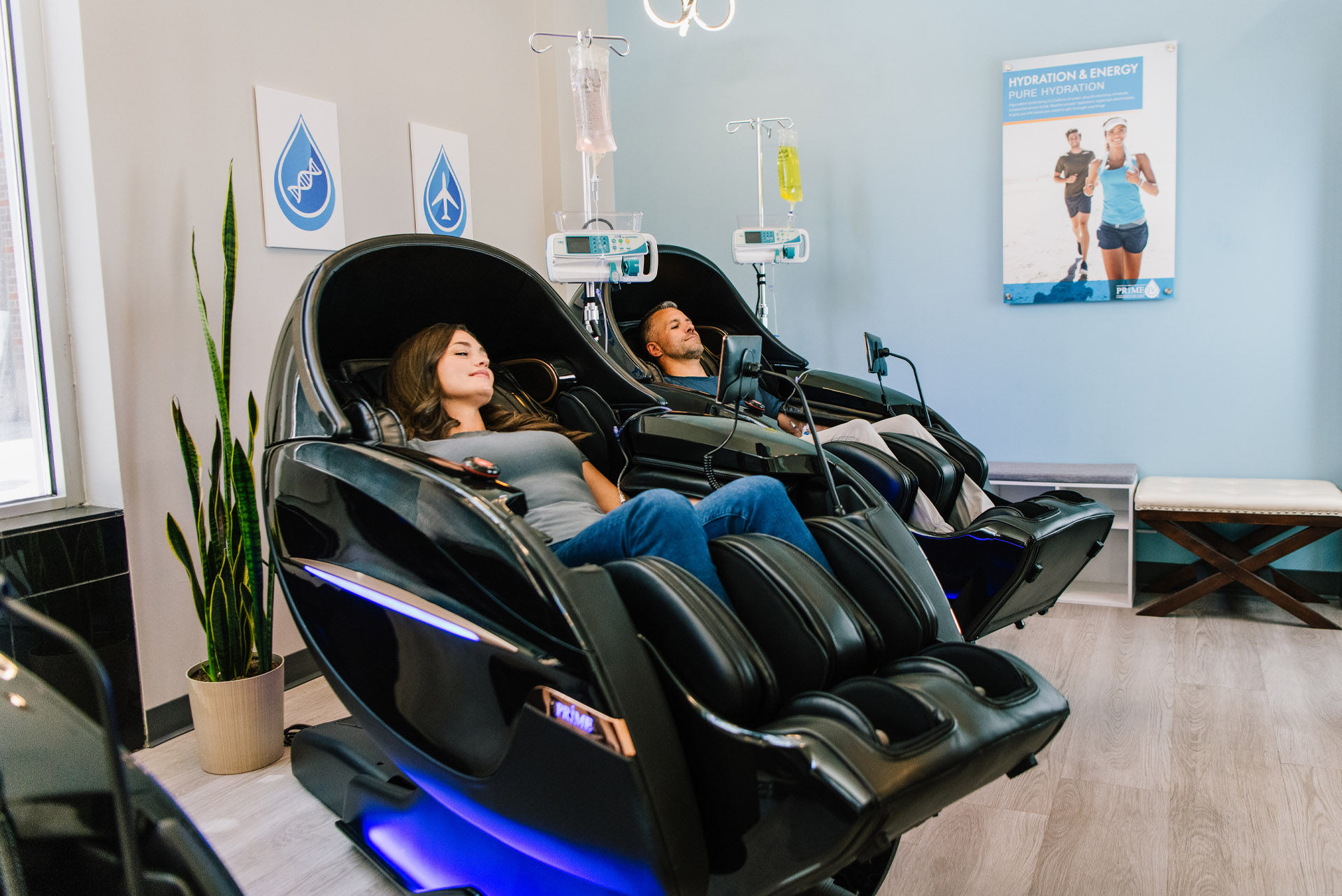 Two adults relaxing in futuristic black massage chairs receiving IV hydration drips in a modern wellness clinic lounge with plant and hydration poster.