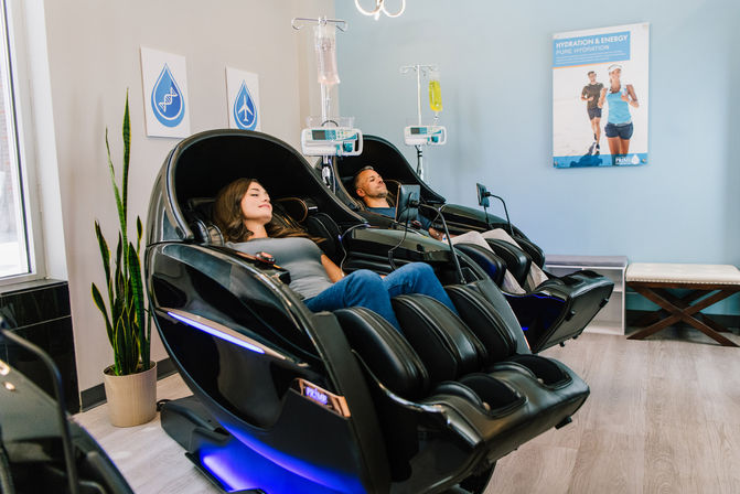 Two adults relaxing in futuristic black massage chairs receiving IV hydration drips in a modern wellness clinic lounge with plant and hydration poster.