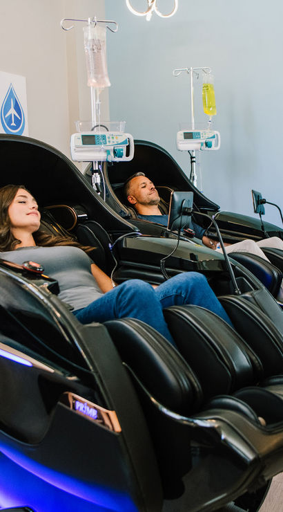 Two adults relaxing in futuristic black massage chairs receiving IV hydration drips in a modern wellness clinic lounge with plant and hydration poster.