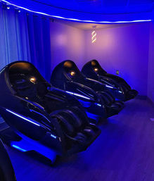 Futuristic glossy black massage chairs lined up in a dim relaxation lounge with blue LED accent lighting, curtains and a spiral pendant lamp