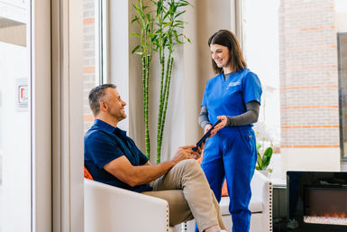 Smiling man seated in a bright healthcare clinic waiting room receives a tablet from a friendly staff member in blue scrubs next to a tall bamboo plant.