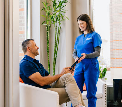 Smiling man seated in a bright healthcare clinic waiting room receives a tablet from a friendly staff member in blue scrubs next to a tall bamboo plant.