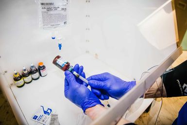 Blue‑gloved hands drawing medication from a brown vial into a syringe inside a sterile clinic prep station, with a 0.9% sodium chloride IV bag and multiple labeled vials visible.