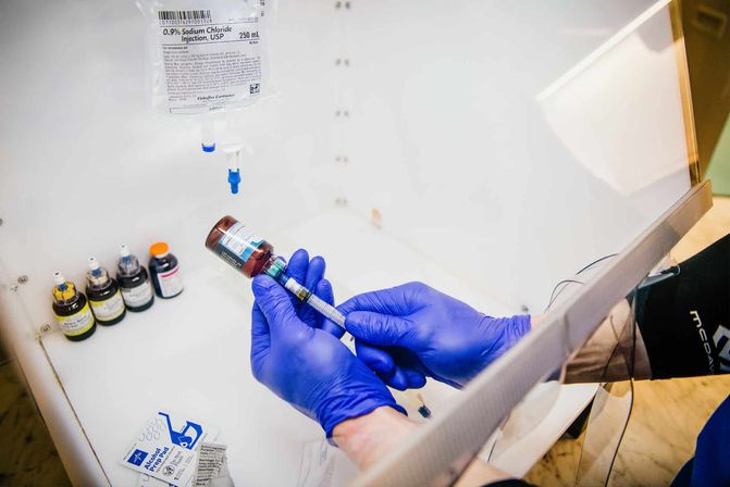 Blue‑gloved hands drawing medication from a brown vial into a syringe inside a sterile clinic prep station, with a 0.9% sodium chloride IV bag and multiple labeled vials visible.