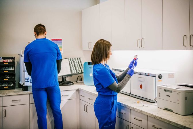 Two healthcare workers in bright blue scrubs in a clean clinic lab — a gloved technician inspects a medication vial at the countertop while a colleague works near storage cabinets and a printer.