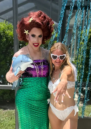 Drag performer in red wig, purple sequin top and green mermaid skirt holding an inflatable dolphin poses with a smiling woman in a white bikini and heart-shaped sunglasses at a sunny backyard poolside party with blue streamers.
