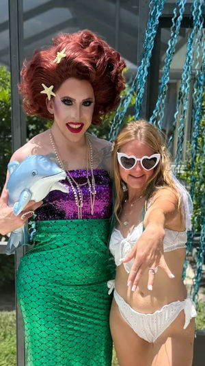 Drag performer in red wig, purple sequin top and green mermaid skirt holding an inflatable dolphin poses with a smiling woman in a white bikini and heart-shaped sunglasses at a sunny backyard poolside party with blue streamers.