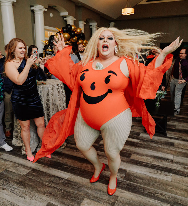 Energetic drag performer in a bright orange smiley-face bodysuit, flowing robe and red heels dancing at an indoor party as guests take photos near black-and-gold balloon decor.