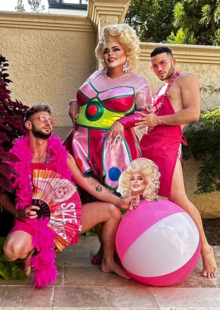 Three people in pink-themed outfits pose on a sunny backyard patio: center drag performer in metallic pink-and-green dress with blonde wig and bold makeup, flanked by two companions in pink accessories and aprons—one with a feather boa and fan, the other wearing pink sunglasses—posed with a large pink-and-white beach ball and cutout face prop.
