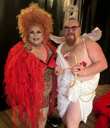 Two costumed performers posing on a stage with a black curtain — a drag artist in a voluminous orange wig, red feathered jacket and leopard-print dress, and a playful cupid in a white toga with wings, heart-shaped sunglasses and a gold bow with heart-tipped arrow.