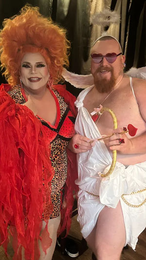 Two costumed performers posing on a stage with a black curtain — a drag artist in a voluminous orange wig, red feathered jacket and leopard-print dress, and a playful cupid in a white toga with wings, heart-shaped sunglasses and a gold bow with heart-tipped arrow.
