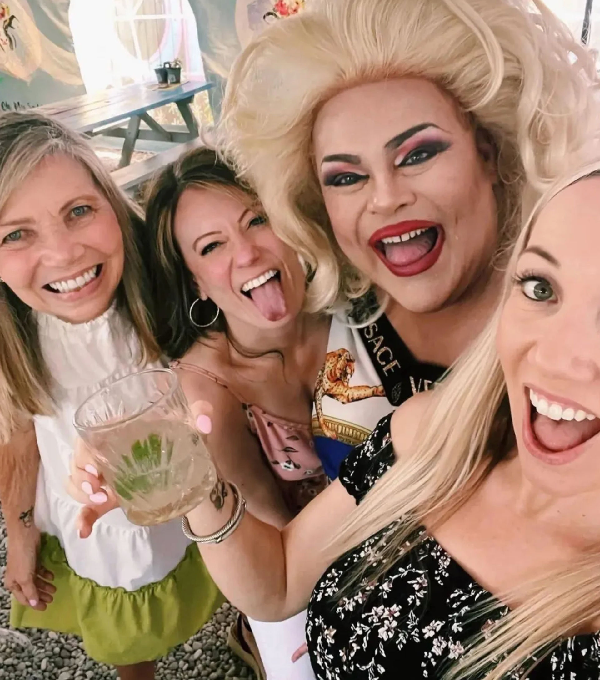 Group selfie of four friends on an outdoor patio bar — smiling women and a drag performer in a blonde wig and bold makeup, one holding a cocktail with lime, lively celebration vibe
