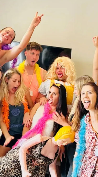 Lively indoor party group of friends wearing colorful feather boas and playful costumes, striking goofy poses and sticking out their tongues.