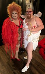 Two costumed performers posing indoors on a wooden floor — a drag artist in a big orange wig, leopard-print dress and red feathered coat, and a playful Cupid in a white toga, wings, heart-shaped sunglasses and toy bow.