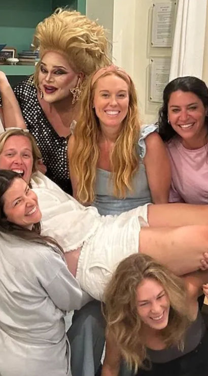 Smiling group of women and a drag performer in a living room, friends playfully lifting one woman horizontally during a cheerful indoor celebration.