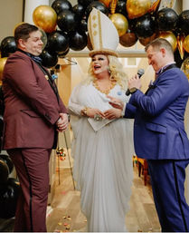 Indoor wedding ceremony with a smiling drag performer officiant in a white gown and tall hat, flanked by two people in burgundy and blue suits under a black-and-gold balloon arch.