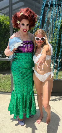 Poolside photo of a drag performer in a green mermaid skirt, purple sequin top and red wig with starfish clips holding a dolphin plush, posing with a smiling woman in a white bikini and heart-shaped sunglasses at a sunny backyard pool party.