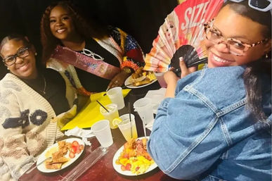 Three smiling friends at a table enjoying a celebratory meal—plates with fried chicken, fruit and drinks—one wearing a glitter sash and another holding a colorful folding fan.