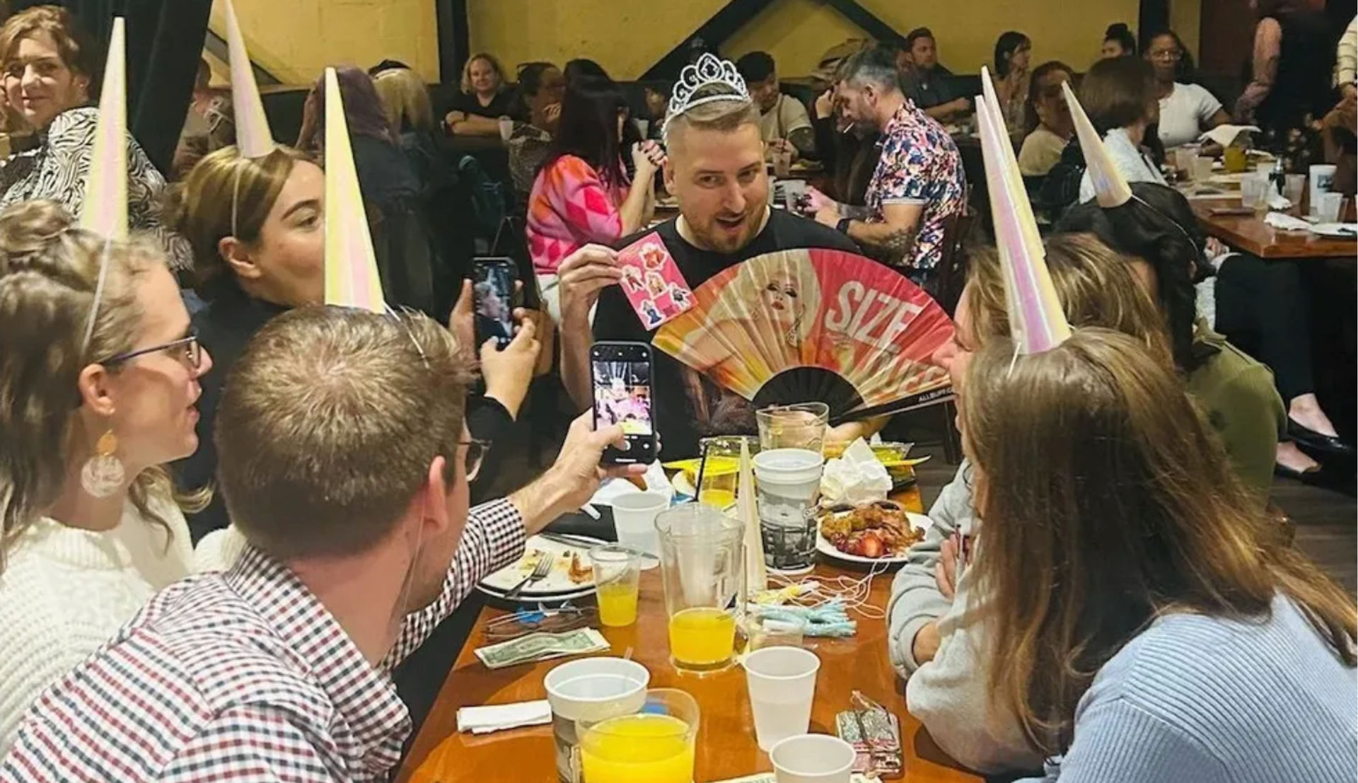 Friends at a lively restaurant celebration wearing pastel cone party hats; a person in a tiara holds a colorful fan and stickers while others snap photos over plates, orange drinks and finger foods.