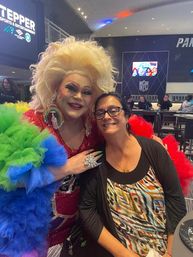 Smiling woman posing with a flamboyant drag performer in a rainbow tulle gown and big blonde wig inside an indoor arena concourse bar area.